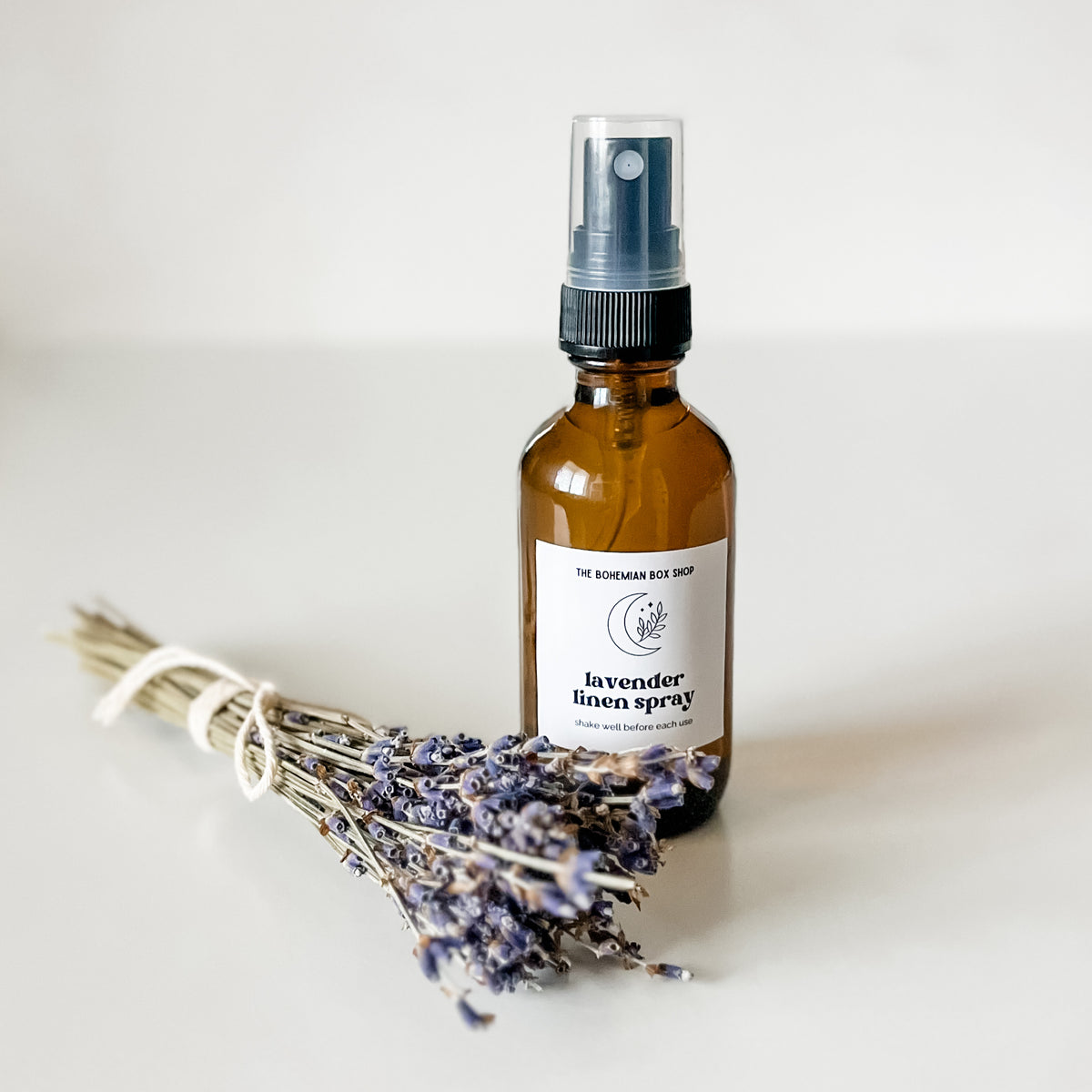 A lavender linen spray 2oz size in an amber spray bottle with a black spray head. Situated on a cream colored background next to a bundle of dried lavender flowers.