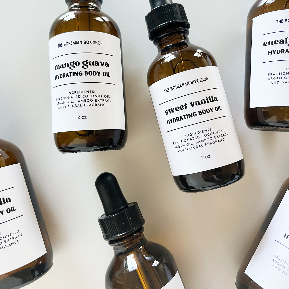 Several amber glass body oil bottles with white labels are arranged on a light neutral background. Visible labels include 'mango guava hydrating body oil' and 'sweet vanilla hydrating body oil' from The Bohemian Box Shop.