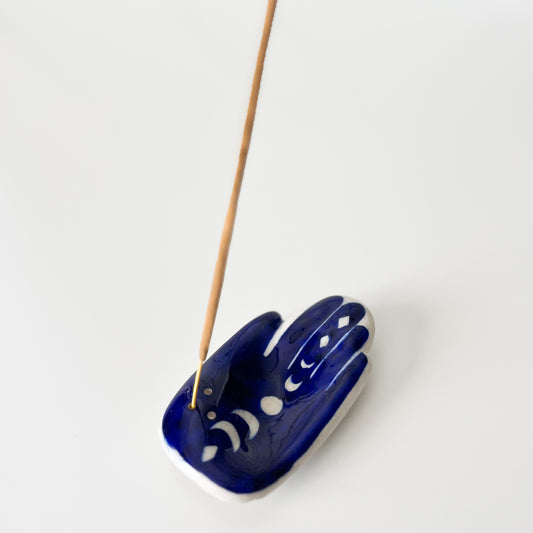 A handcrafted ceramic incense holder in the shape of a Hamsa hand with a moon phase design, painted in blue color, with an incense stick inserted.