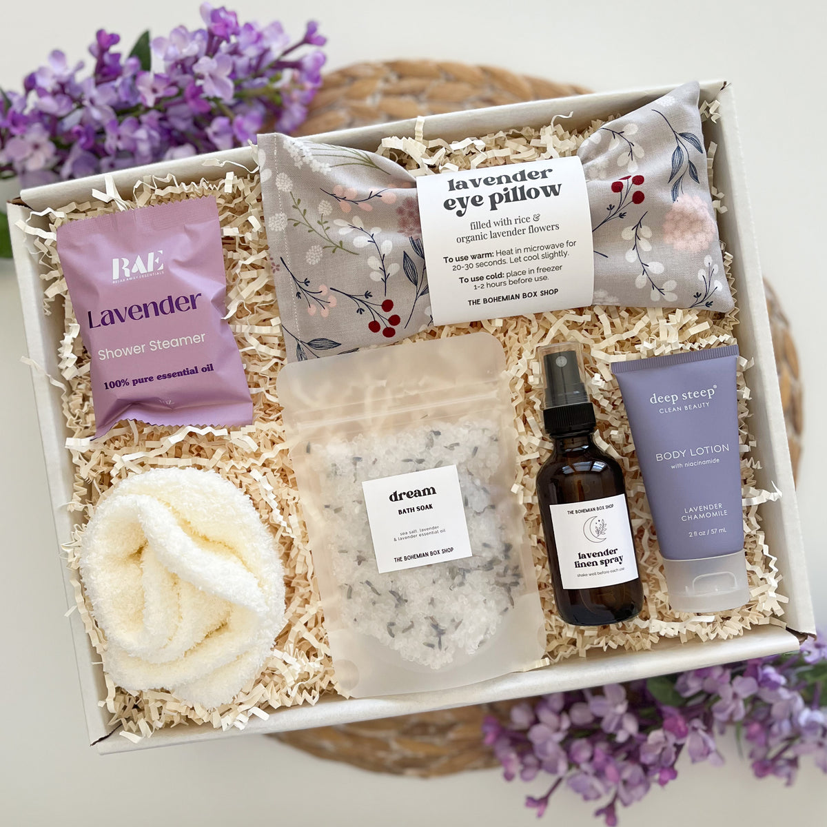 A curated spa gift box made by The Bohemian Box Shop, containing a lavender shower steamer packet, a lavender eye pillow, women's white cozy socks, a lavender bath salt packet, a lavender linen spray bottle, and a lavender body lotion tube, surrounded by purple lavender flowers.