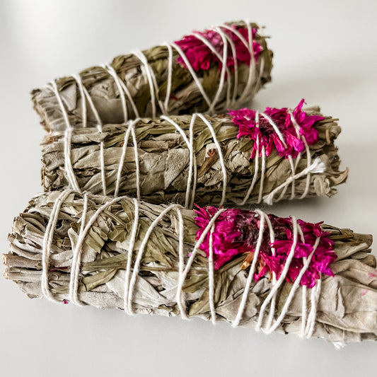 White sage smudge sticks with pink dahlia flowers tied to the top, arranged in a spiral on a light background.
