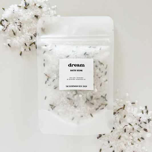 A sealed clear plastic packet labeled 'dream' containing white bath soak packets against a white background, with decorative lavender flowers scattered around.
