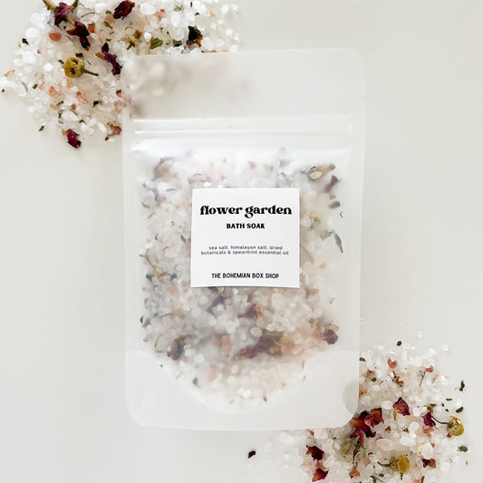 A clear packet labeled 'Flower Garden Bath Soak' with a mix of dried botanicals and essential oils, displayed on a white surface with decorative flowers around it.