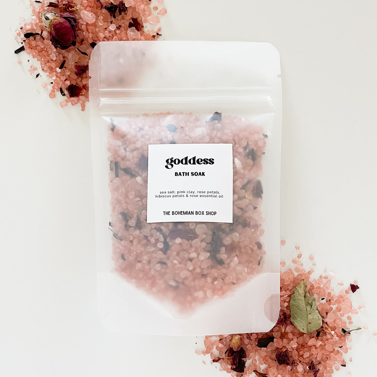 A clear packet of bath soak with rose and hibiscus, surrounded by pink rose petals and a few leaves on a white background. The label reads 'goddess bath soak' and it's made by The Bohemian Box Shop