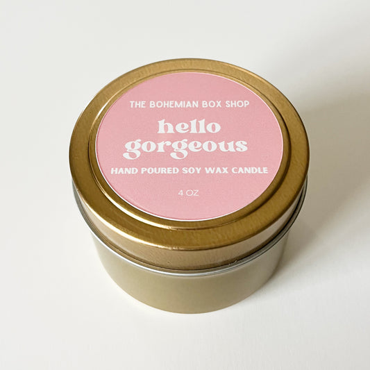Candle in a gold tin with a pink label that says 'hello gorgeous' on a white background