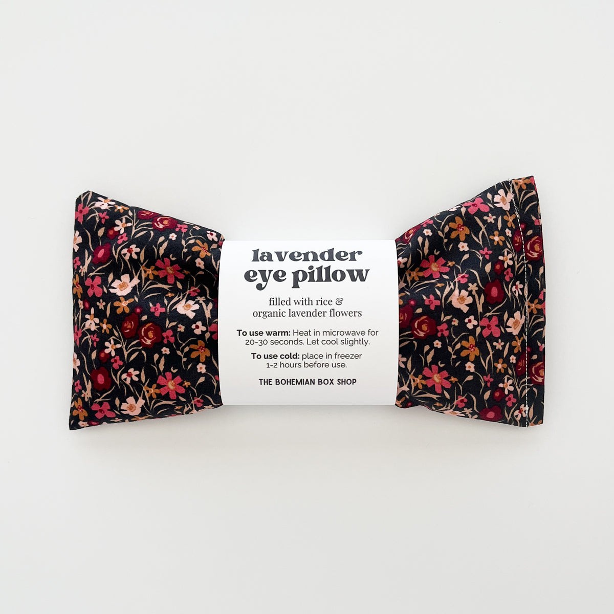 A lavender eye pillow by The Boheman Box Shop, featuring a black fabric base with a red, pink, and orange floral pattern on both sides, wrapped in a white paper label in the center.