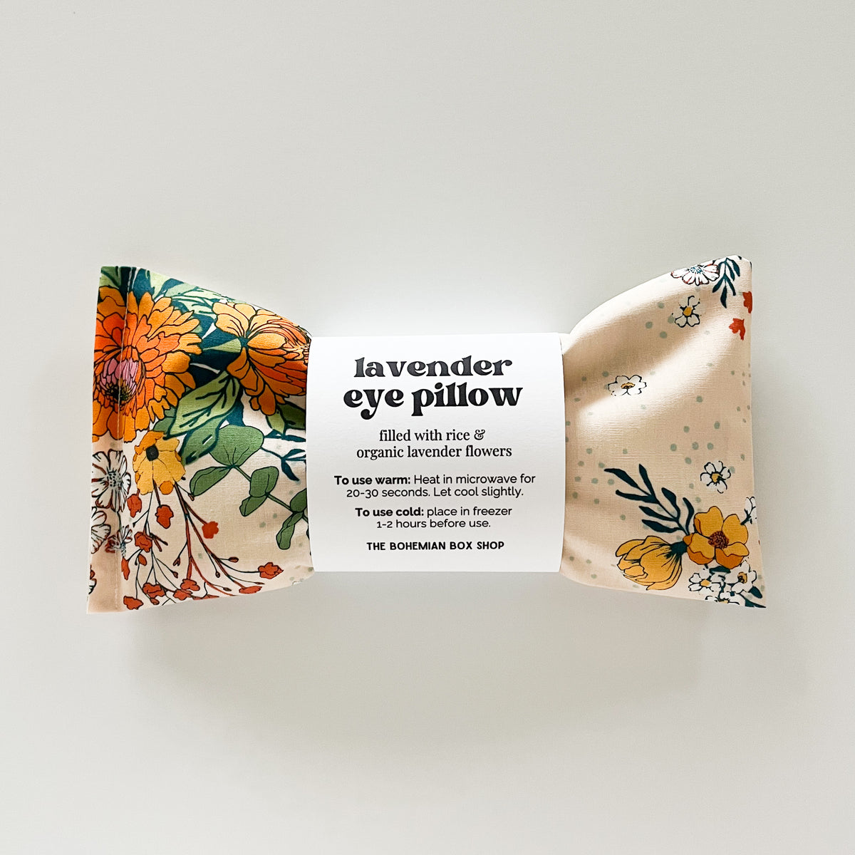 A lavender eye pillow by The Bohemian Box Shop, in a marigold floral-patterned fabric with a white paper label in the center.