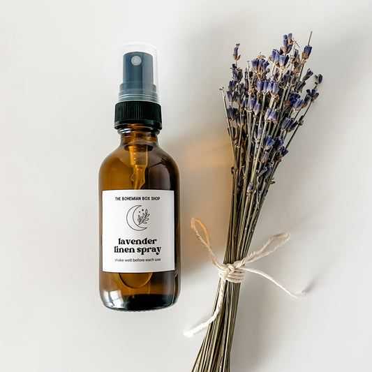 A 2oz amber glass bottle of lavender linen spray with a black spray nozzle, labeled 'lavender linen spray'. Accompanied by a bundle of dried lavender tied with a string, all placed on a white surface.