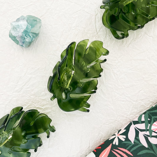 Three green hair claws with a monstera leaf design, displayed on a white surface next to a crystal and a floral patterned fabric.
