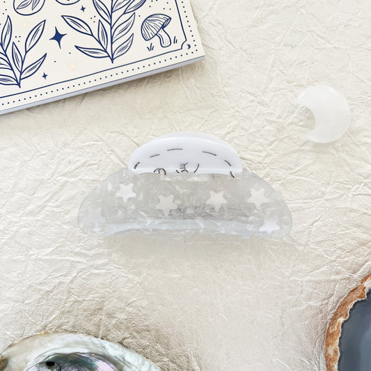 A silver hair claw with moon and stars design, placed on a textured surface next to a hair clip and a note pad.