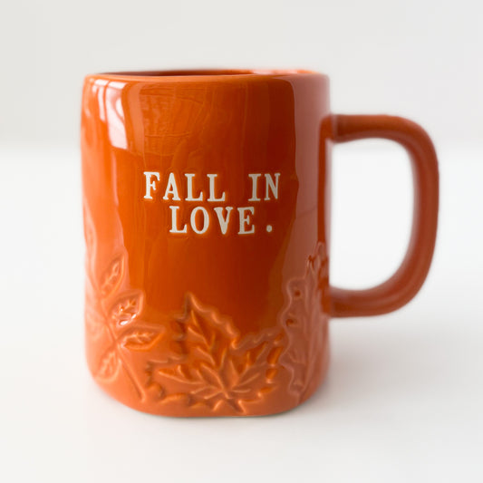 An orange ceramic mug with embossed autumn leaf details and the phrase 'FALL IN LOVE.' printed in white on the front, set against a plain white background.