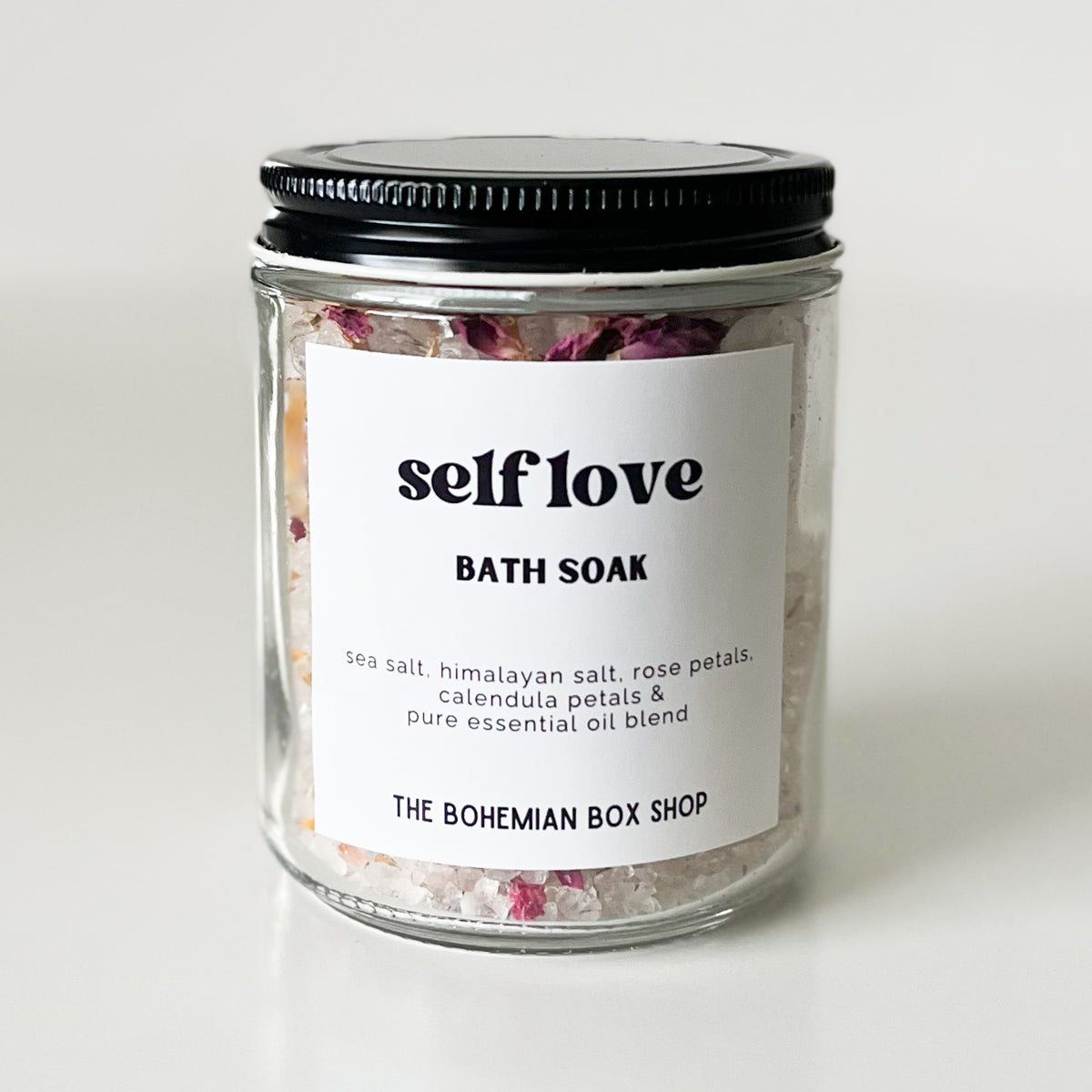 A glass jar of bath soak with a black metal lid and a white label reading 'self love BATH SOAK' and 'THE BOHEMIAN BOX SHOP'. The jar contains white bath salts with visible pink and purple dried flower petals.
