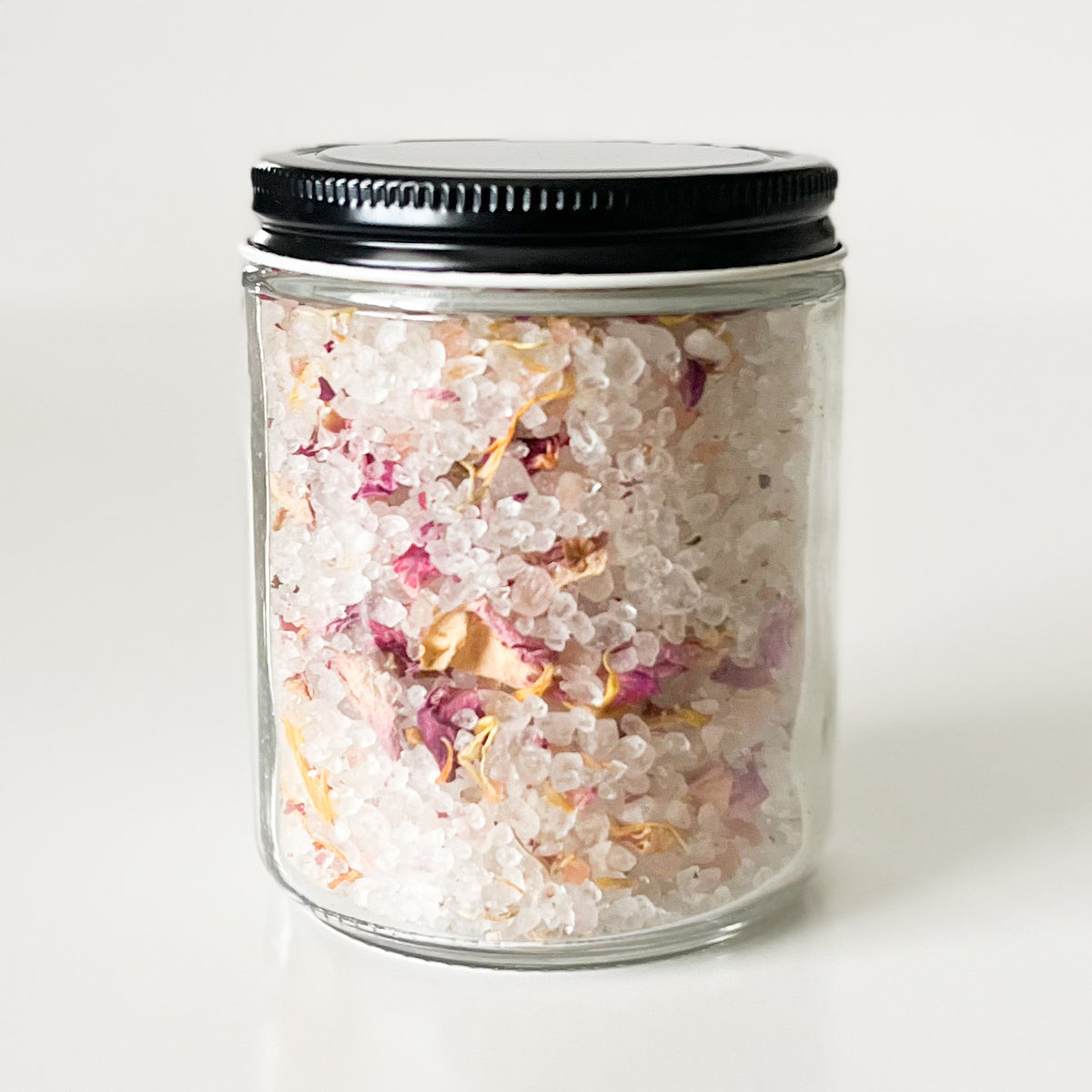 A clear glass jar filled with white bath salts mixed with dried pink and yellow flower petals, sealed with a black metal lid, on a clean white background.