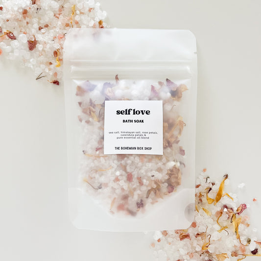 A frosted clear packet labeled 'Self Love Bath Soak' by the Bohemian Box Shop with a mix of rose and calendula petals visible through the packet, placed on a white surface with more petals scattered around.