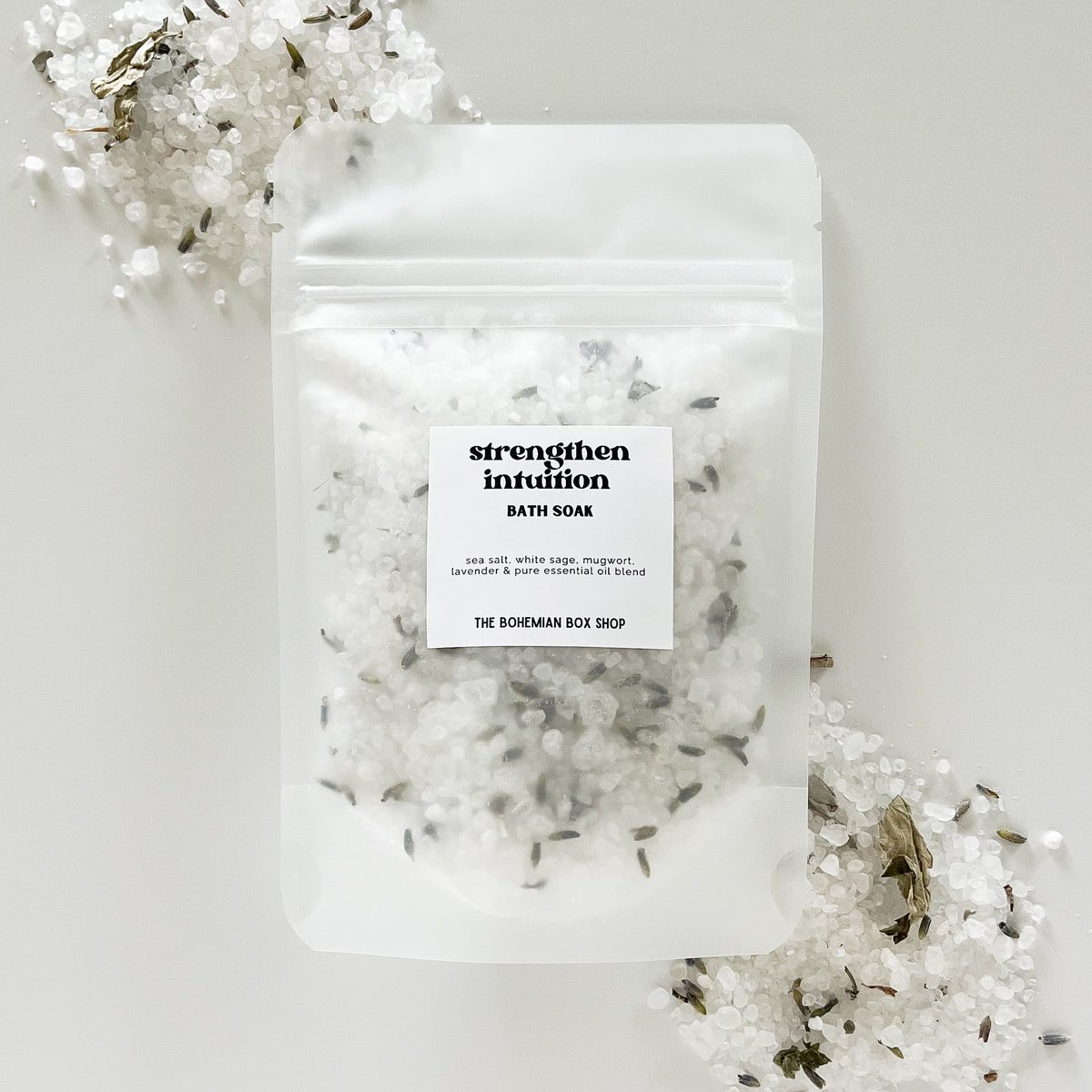 A packet of 'Strengthen Intuition Bath Soak' by The Bohemian Box Shop placed on a white surface surrounded by dried white sage leaves.