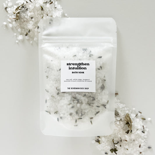 A packet of 'Strengthen Intuition Bath Soak' by The Bohemian Box Shop placed on a white surface surrounded by dried white sage leaves.