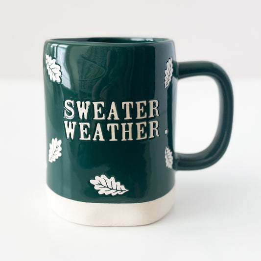 A dark green ceramic mug with a cream-colored base and handle, featuring the text 'SWEATER WEATHER' and small white leaf illustrations on a clean white background.