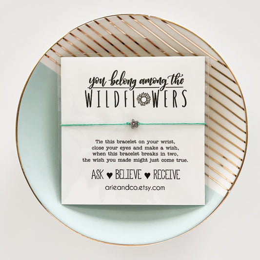 A wish bracelet with a card that reads 'You Belong Among The Wildflowers' and has a small flower bead, presented on a turquoise plate with a geometric pattern.