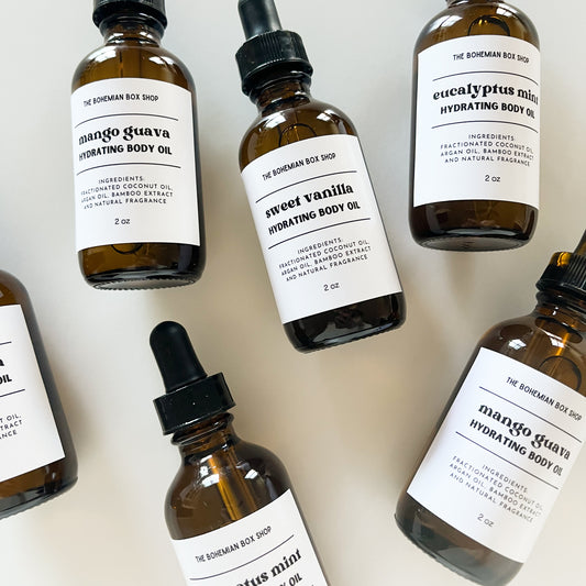 Six amber glass dropper bottles of hydrating body oil arranged on a light beige background. The bottles have white labels with black text and are topped with black droppers 