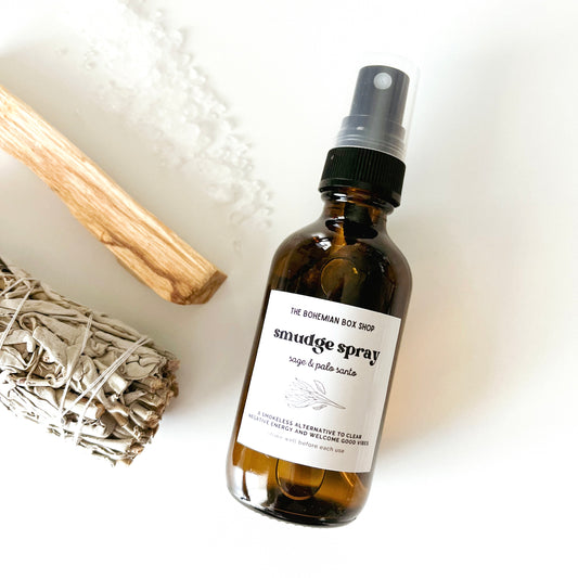A brown bottle labeled 'smudge spray' with a spray nozzle, accompanied by a bundle of Palo Santo wood and White Sage leaves on a light background.