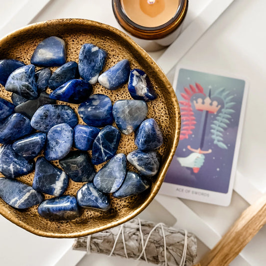 Sodalite Tumbled Stones on a plate with a tarot card, white sage bundle, palo santo stick and candle in the background