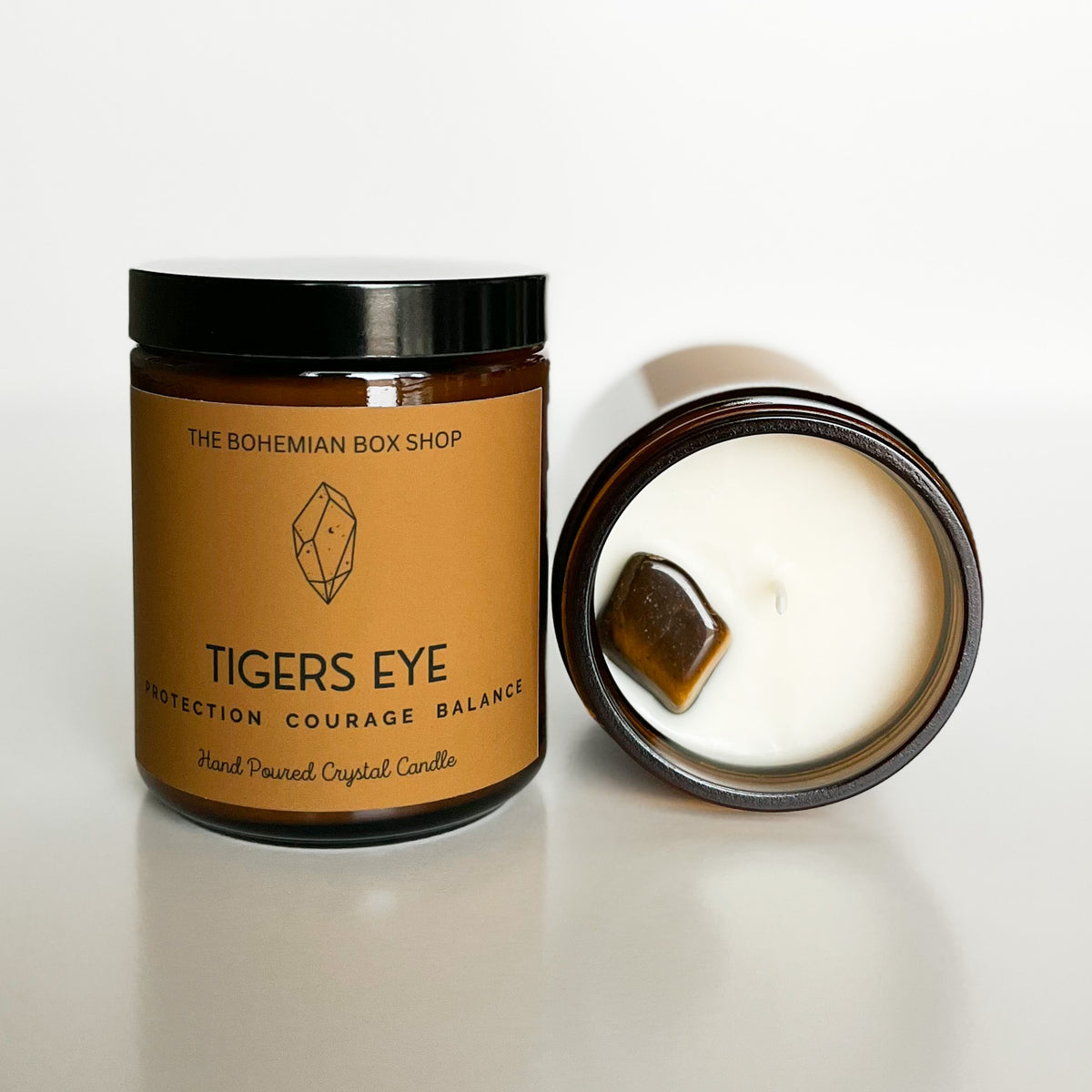 A brown soy candle with a label reading 'Tigers Eye' and 'The Bohemian Box Shop', alongside a candle showing the melted wax and a piece of Tigers Eye crystal inside.